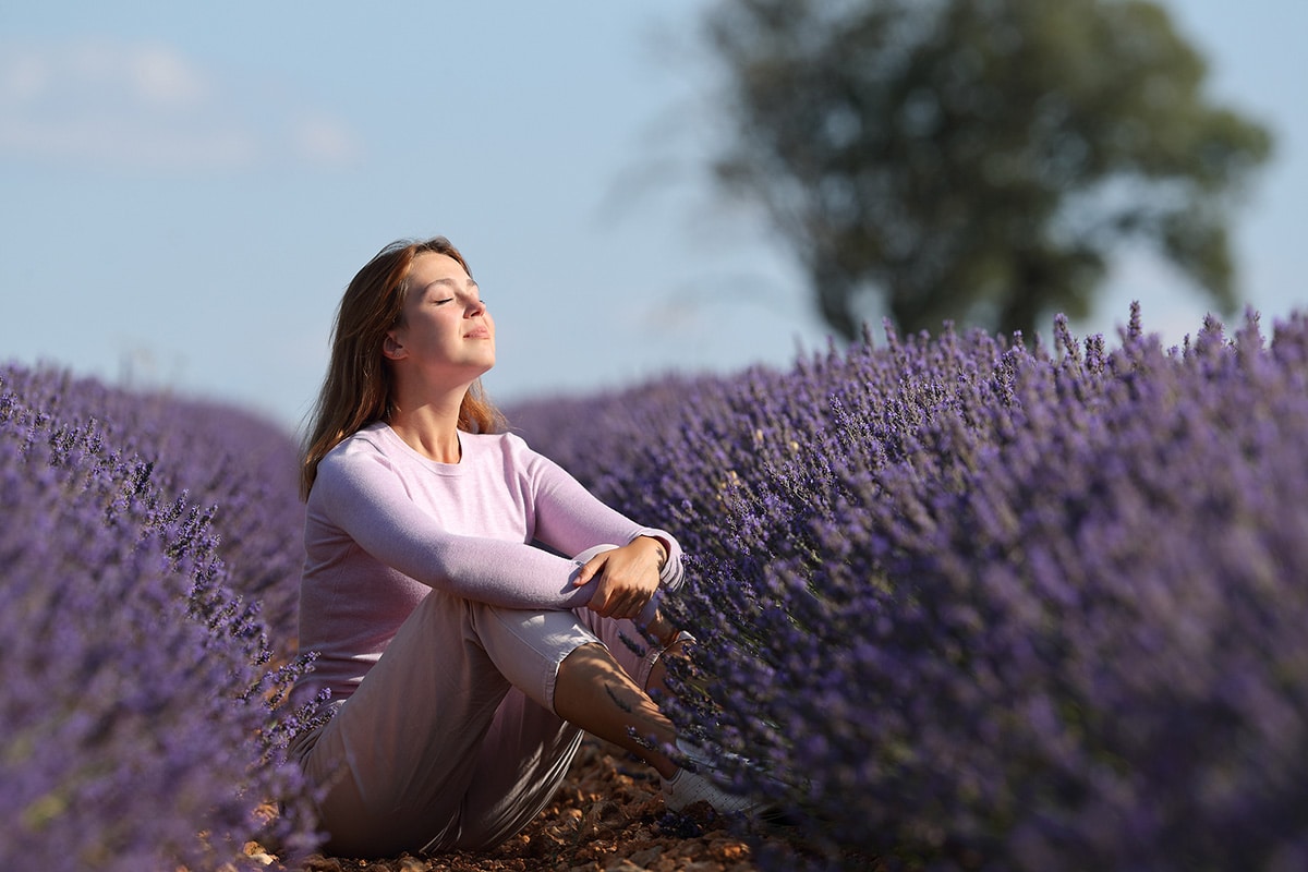 Woman sitting in field of lavender with eyes closed and face lifted to the sun.