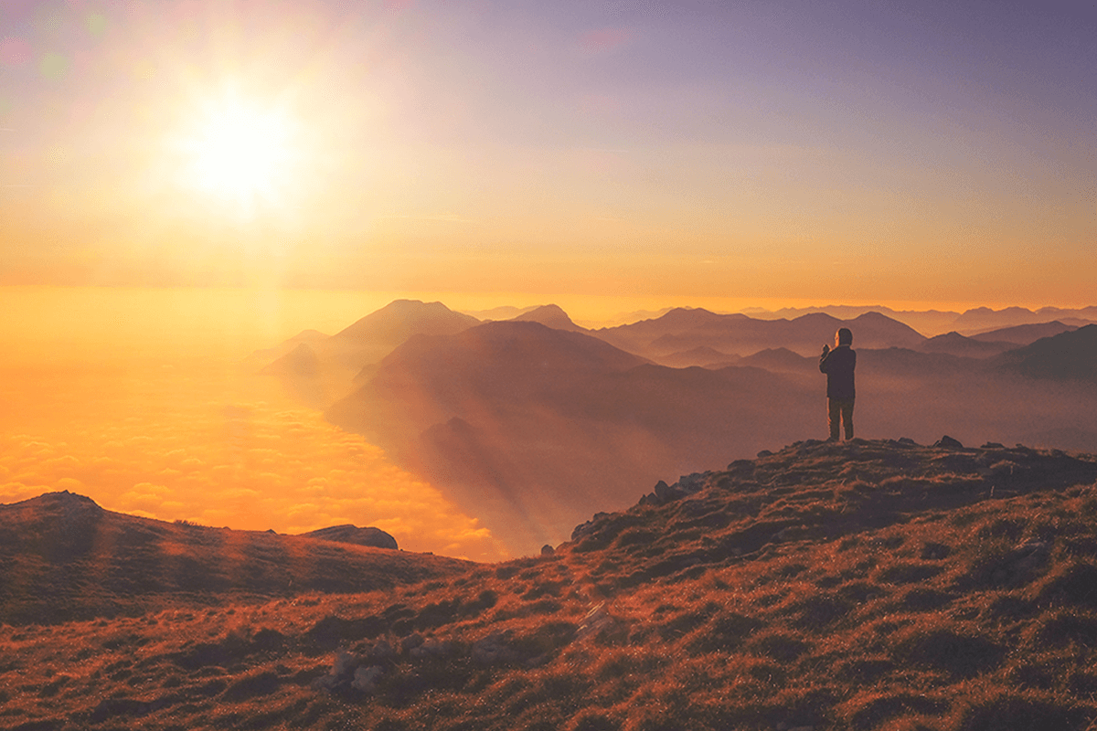 Person standing on hillside looking out over mountains with sun setting in the distance