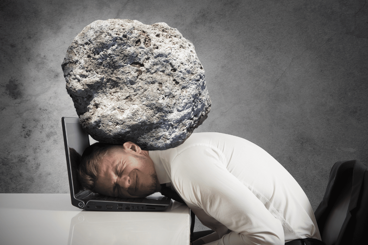 Man at desk with head on computer, neck getting crushed by a rock to illustrate feeling stressed.